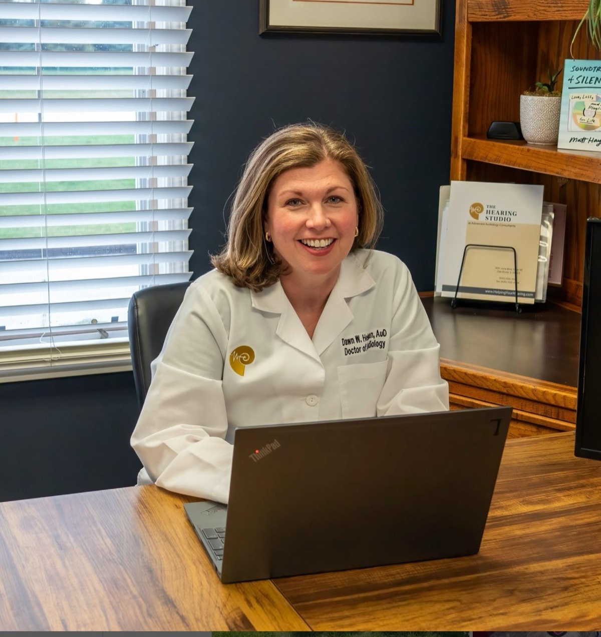 Dr. Dawn Heiman, AuD, in her white clinic coat, smiling from behind the laptop at the desk in her audiology practice.